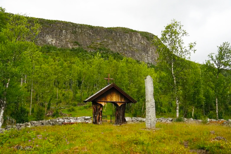 Bilde av en treportal med steingjernde og en bauta. I Bakgrunnen er skog og et berg. 