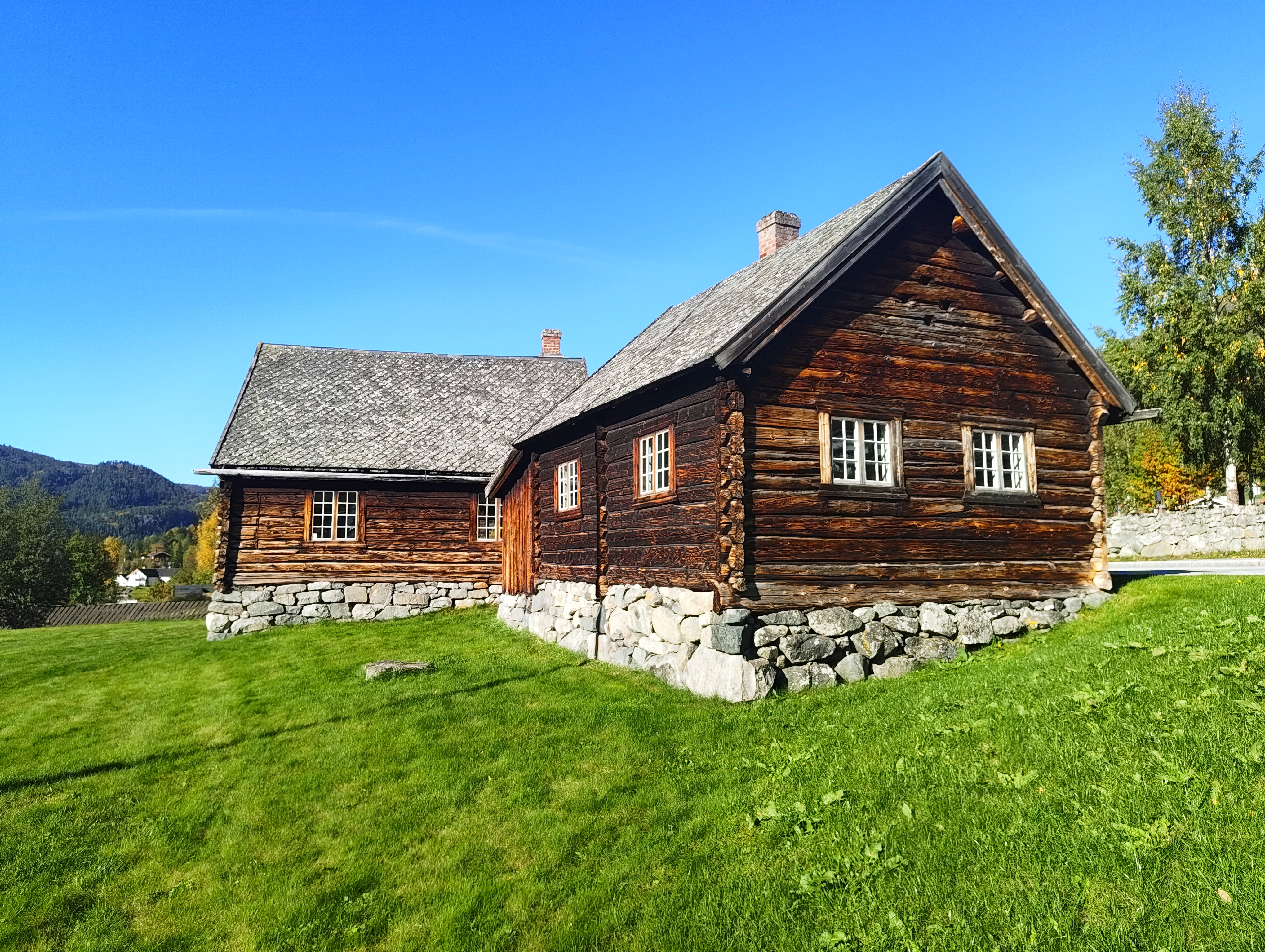 Bilde av to gamle lafta bygninger med gressplen rundt på en solfylt sommerdag. 