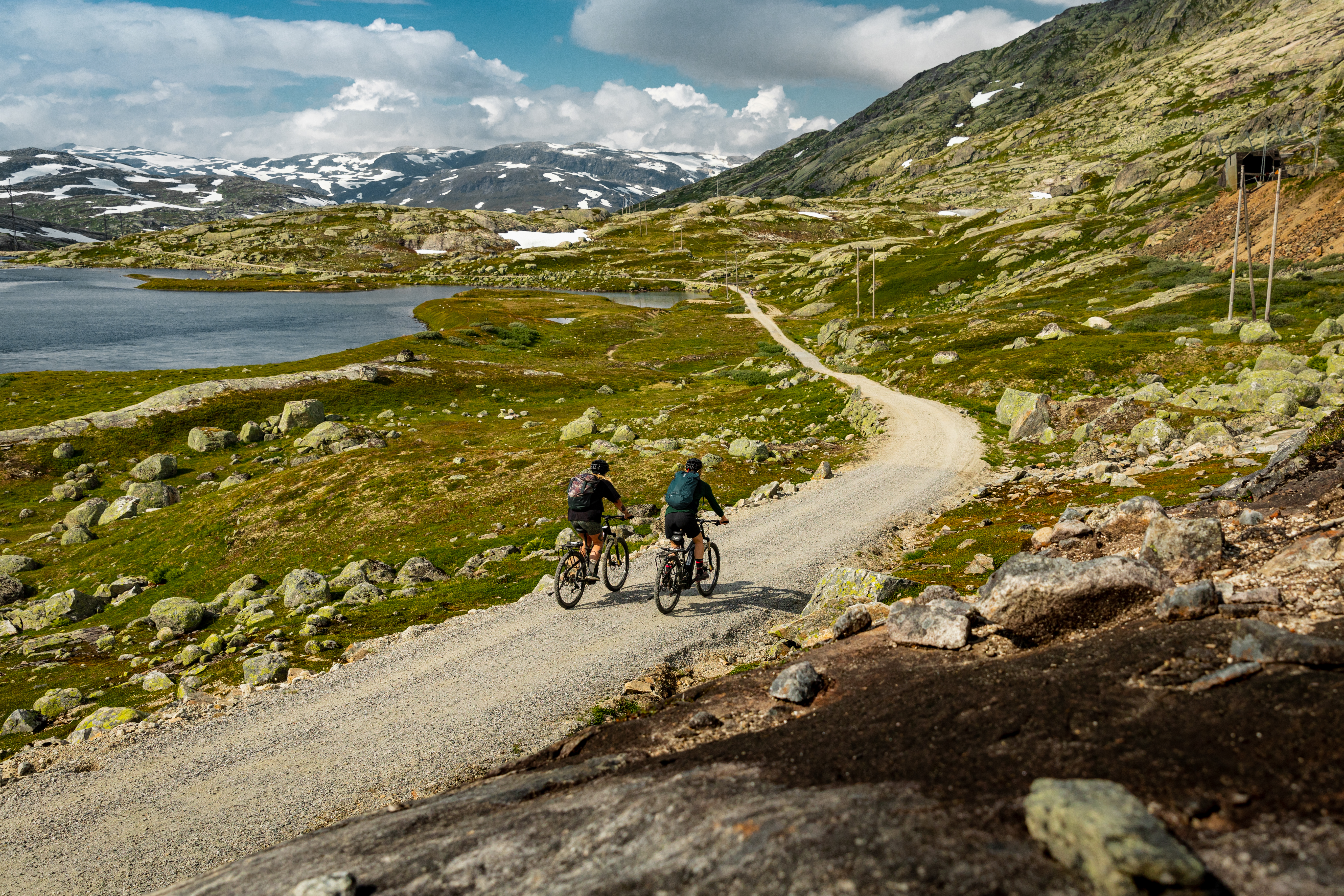 Bilde av to syklister på en grusvei på høyfjellet. Det er et vann og fjellandskap i bakgrunnen. 