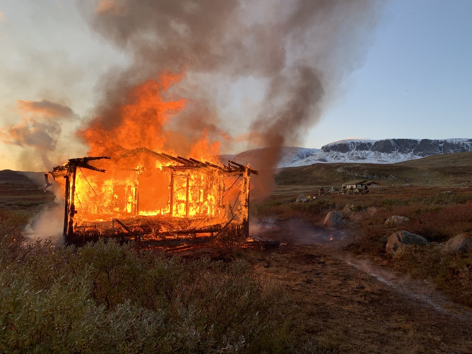 Foto av et hus i full fyr på fjellet, med solnedgang i bakgrunnen.