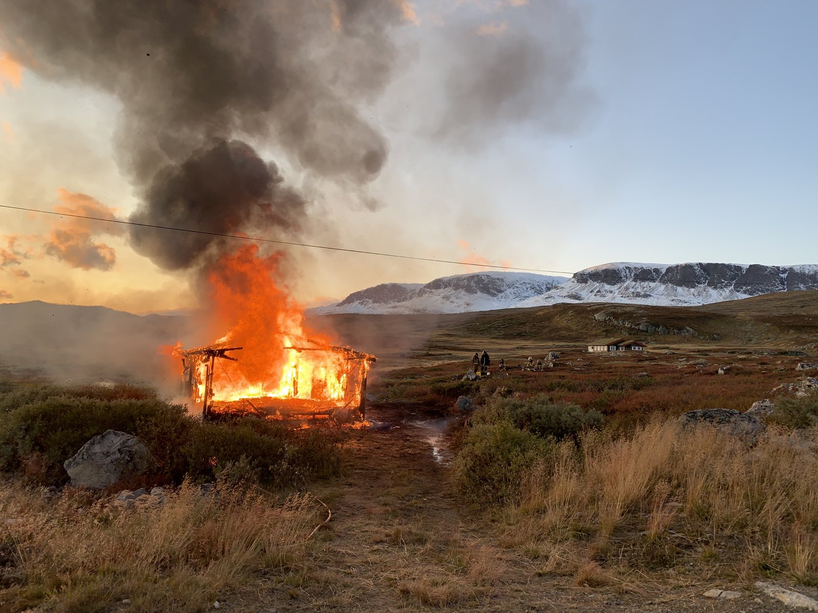 Bilde av hytte som brenner på fjellet. Snøkledd skarv i bakgrunnen. 